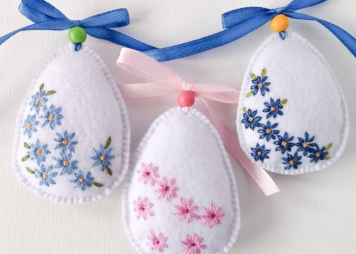 Easter eggs with daisy embroidery. White DIY felt ornaments with blue and pink embroidered flowers