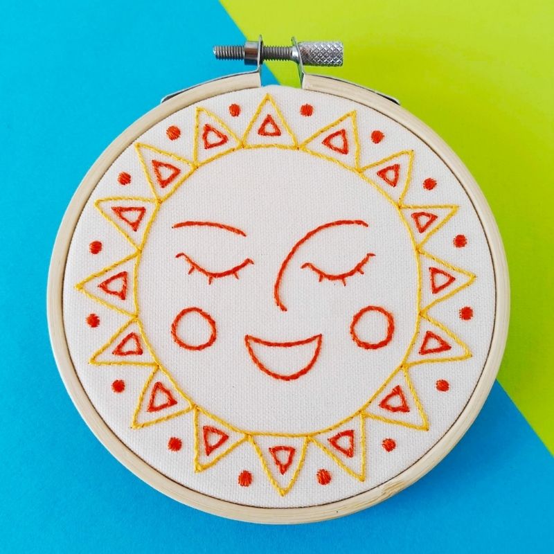 Smiling sun face hand embroidered with yellow and orange threads on white linen, framed in a bamboo hoop