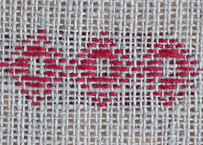 A darning stitch in red thread in the shape of a diamond on loose woven fabric courtesy of practicalembroidery.eu