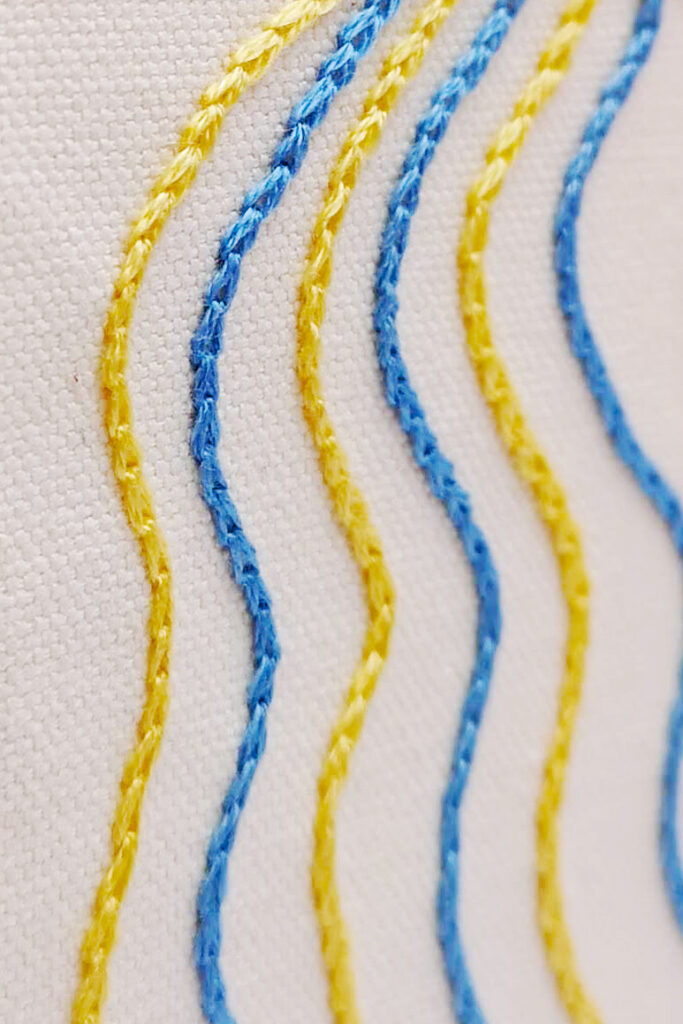 Handgestickte Kettenstiche in gelben und blauen Farben auf weißer Leinwand.